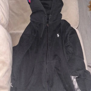 Svart hoodie från Polo Ralph Lauren - Svart hoodie från Polo Ralph Lauren med dragkedja och klassisk vit logga på bröstet. Tröjan har huva med snörning och två fickor framtill. Tillverkad i mjuk bomull, perfekt för en chill och stilren look. Helt ny i skicket också