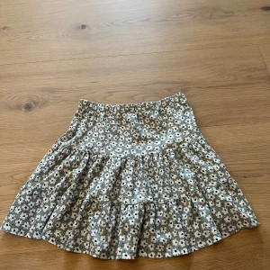 Blommig volangkjol från 157.New - Supersöt kort volangkjol från 157. Med blommigt mönster i vitt och svart på en ljusgrön bakgrund. Kjolen har resår i midjan och är gjord i ett mjukt, lätt material som känns skönt mot huden. Perfekt för dig som gillar en chill och trendig stil.