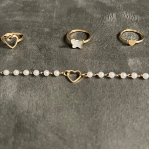 Guldiga ringar & pärlarmband med hjärta - Säljer ett set med tre tunna guldiga ringar, en med hjärta, en med vit fjäril och en med litet hjärta. Till det hör ett armband med vita pärlor och ett hjärta i guld. Perfekt för dig som gillar söta detaljer och stilren design.
