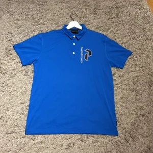 Blå pikétröja Peak Performance XL - Snygg blå pikétröja från Peak Performance i storlek XL. Tröjan har klassisk krage, tre knappar och logga broderad på bröstet. Perfekt för dig som gillar sportig och stilren look. Tillverkad i mjukt och lätt material som andas.
