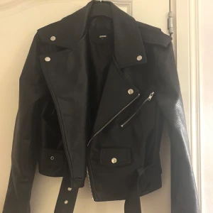 Svart skinnjacka från Bik Bok - Snygg svart skinnjacka från Bik Bok med klassisk bikerstil. Jackan har silvriga detaljer, dragkedjor, bälte i midjan och breda slag. Perfekt för att lyfta vilken outfit som helst och ge en edgy vibe.
