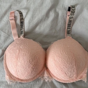 Rosa spets-bh från Victoria's Secret - Supersöt ljusrosa bh från Victoria's Secret med blommig spets och vadderade kupor. Axelbanden är dekorerade med glittriga stenar och text. Perfekt för dig som vill ha något extra snyggt under kläderna.