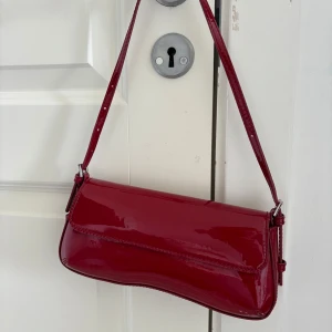 Röd lackad axelväska i retrostil - Säljer en snygg röd axelväska med glansig lackad yta och stilren design. Väskan har en smal axelrem och en mjukt böjd form, perfekt för att lyfta vilken outfit som helst. Enkel att matcha och ger en cool retrovibe.