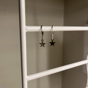 Silvriga örhängen med stjärnhänge - Rostfritt stål.Säljer ett par stilrena silvriga örhängen med små stjärnhängen. Perfekta för dig som gillar minimalistiska smycken med en cool touch. Passar till alla tillfällen och är lätta att matcha med andra accessoarer. Handgjort 