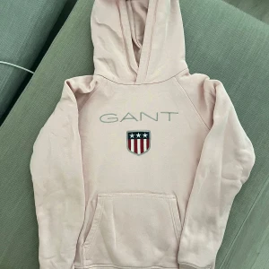 Ljusrosa hoodie från GANT med logga - Säljer en ljusrosa hoodie från GANT med broderad logga och sköld på bröstet. Tröjan har huva, magficka och långa ärmar. Mjuk insida och klassisk passform, perfekt för dig som gillar stilren och sportig look.