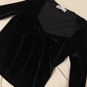 Svart sammetsblus från Hollister - Supersnygg svart sammetsblus från Hollister med lång ärm och en söt knutdetalj framtill. Toppen har en figurnära passform och en djupare urringning som ger en trendig look. Perfekt att styla med jeans eller kjol för en cool outfit.