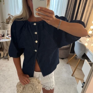 Mörkblå jeansjacka med puffärm - Säljer en mörkblå jeansblus med puffiga trekvartsärmar och silverfärgade knappar framtill. Jackan har en loose fit och är tillverkad i klassiskt denimtyg. Perfekt för dig som vill ha en chill och trendig look. Aldrig använd!