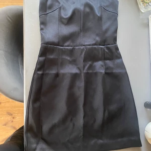 Svart axelbandslös kort klänning - Snygg svart axelbandslös klänning i kort modell med figurnära passform. Klänningen har en slät yta med lätt glans och markerad midja, perfekt för en stilren look. Passar dig som vill ha en enkel men elegant outfit.