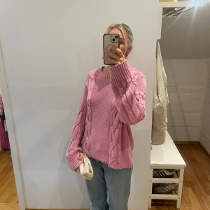 Rosa stickad kabeltröja - Mysig rosa stickad tröja med grova kabelstickade detaljer längs ärmar och fram. Tröjan har rund halsringning och långa ärmar med breda muddar. Perfekt för att addera lite färg till din höst- eller vintergarderob.