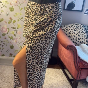 Leopardmönstrad långkjol med slits - Snygg långkjol med leopardmönster i beige och svart. Kjolen har en rak passform och en hög slits på sidan som ger en cool vibe. Perfekt att styla med boots eller sneakers för en trendig look.