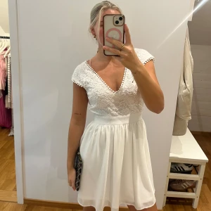 Vit spetsklänning med v-ringning - Superfin vit kortklänning med spets upptill och v-ringad hals. Kjolen är lätt och flowy, och klänningen har korta ärmar. Perfekt för sommarens alla tillfällen. Snygg passform och feminina detaljer.