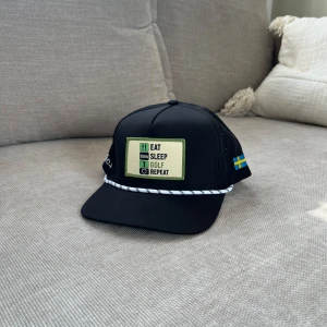 Svart keps Golfio.se Eat Sleep Golf - Svart keps med böjd skärm, perforerade sidor och justerbar snapback. Framtill sitter ett broderat märke med texten 'Eat Sleep Golf Repeat' i grönt och beige. Sidan har svensk flagga och Golfie.so-brodyr. Vit och svart snodd framtill ger extra stil.
