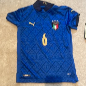 Italien  fotbollströja med Veratti #6 på ryggen - Blå Italien fotbollströja från Puma med guldiga detaljer, nummer 6 och Veratti på ryggen. Tröjan har korta ärmar, mönstrad framsida och Italiens landslagsemblem på bröstet. Tröjan är i väldigt bra skick