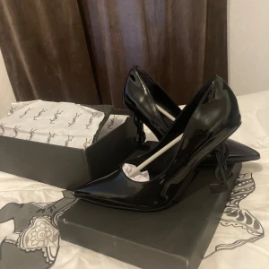 Svarta Saint Laurent pumps med logoklack - Säljer ett par svarta pumps från Saint Laurent i lackat skinn med spetsig tå och hög klack formad som YSL-loggan. Skorna har en elegant och edgy vibe, perfekta för att sticka ut. Kommer med originalkartong och dustbag.