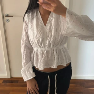 Vit blus med broderade detaljer - Superfin vit blus med broderade hålmönster och v-ringning. Blusen har långärmade, lite vida ärmar och markerad midja som ger en snygg siluett. Perfekt för vår och sommar när du vill ha en luftig och söt topp med romantiska vibbar.