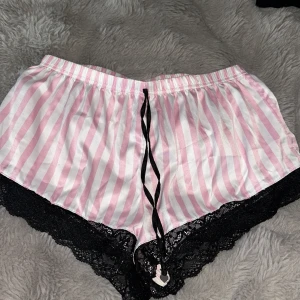 Randiga pyjamasshorts med spets från Censored - Söta pyjamasshorts från Censored i rosa och vitt med vertikala ränder. Shortsen har svart spetskant nertill och en svart snörning framtill. Tillverkade i ett mjukt, glansigt material som känns lyxigt mot huden. Perfekta för mysiga hemmakvällar.