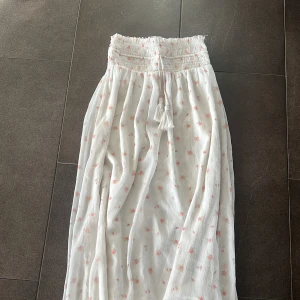 Vit blommig långkjol från Gina Tricot - Supersöt långkjol från Gina Tricot i vitt med små rosa blommor. Kjolen har smock i midjan, dekorativa snören med tofsar och är gjord i ett lätt, luftigt tyg. Perfekt för sommaren!