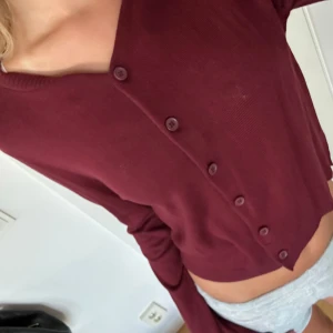 Vinröd  kofta med knappar - Säljer en vinröd  kofta med långa ärmar och knappar framtill. Tröjan har en enkel, stilren design och är tillverkad i ett mjukt, stickat material som känns skönt mot huden. Aldrig andvänd.