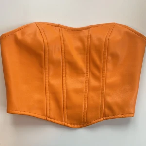 Orange korsettopp i skinnimitation - Snygg orange korsettopp i skinnimitation med markerade sömmar och figurnära passform. Toppen är axelbandslös och har dragkedja i sidan för enkel på- och avtagning. Perfekt för dig som vill sticka ut med en cool och trendig look. Använd 1 gång! Storlek XS. 🧡