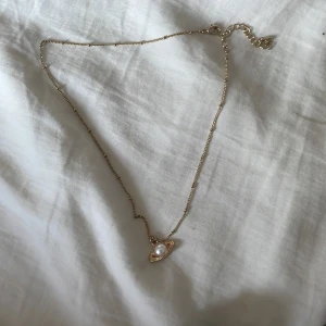 Guldigt halsband med planet-hänge - Snyggt halsband i guld med ett unikt planetformat hänge. Hänget har en vit pärla i mitten och små glittriga detaljer runt om. Perfekt accessoar för dig som gillar rymdinspirerade smycken och vill sticka ut lite extra.