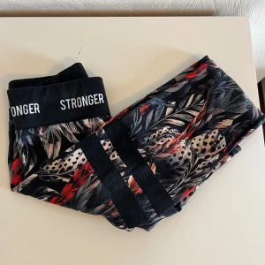 Mönstrade leggings från Stronger - Snygga leggings från Stronger med svart bas och färgglatt fjädermönster i rött, beige och grått. Bred resår i midjan med logga och två svarta ränder på ena benet. Väldigt bra skick men använda. 