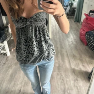 Leopardmönstrad bandeau topp - Säljer en bandeau topp med leopardmönster i svart och grått. Toppen är ärmlös och har en lätt rynkad detalj under bysten som ger en snygg passform. Perfekt att styla med jeans eller kjol för en cool look går också att ha som en miniklänning