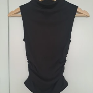 Svart ribbad polotopp med rynkade sidor - Snygg svart ärmlös polotopp med ribbad struktur och rynkade detaljer längs sidorna. Toppen har en tight passform och hög hals, perfekt att styla med jeans eller kjol för en clean look.