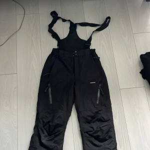 Svarta täckbyxor Base Camp S - Svarta täckbyxor från Base Camp med hängslen och dragkedjor på fickorna. Byxorna har hög midja och är gjorda i ett slitstarkt syntetmaterial, perfekta för vintern. Snygg och funktionell design med justerbar midja och förstärkta benslut.