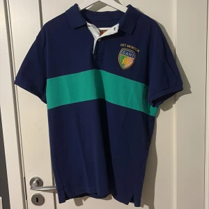 Blågrön pikétröja från Gant - Snygg marinblå pikétröja från Gant med grön bred rand över bröstet och ryggen. Klassisk krage, korta ärmar och broderad Gant-logga på vänster bröst. Tillverkad i mjuk bomull, perfekt för en sportig och stilren look.