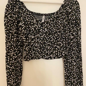 Blommig svart blus  - Supersöt svart blus med vita och gula små blommor över hela tyget. Blusen har lång ärm, v-ringning och knappar framtill. 