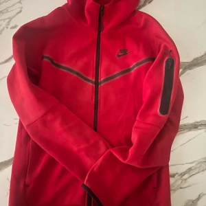 Röd Nike vindjacka med huva - Nike vindjacka i rött med svarta detaljer och dragkedja hela vägen. Jackan har huva, lång ärm och en svart rand över bröstet. Praktisk ficka med dragkedja på ena ärmen och klassisk Nike-logga på bröstet. Perfekt för dig som gillar sportig stil.