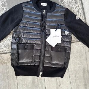 Svart Moncler pufferjacka med dragkedja - HELT NY! Snygg svart pufferjacka från Moncler med quiltad framsida, två stora fickor med knappar och ribbade muddar. Jackan har långärmade ärmar i mjukt tyg och klassisk Moncler-logga på ärmen. Perfekt för dig som vill ha en stilren och exklusiv look.