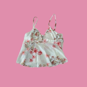 Blommig vit linnetopp med volang - Supersöt vit linnetopp med rosa och röda blommor, tunna axelband och volangdetalj under bysten. Luftig och flowy passform, perfekt för varma dagar. Ger en romantisk och somrig vibe.