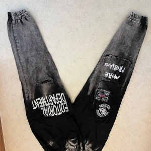 Svarta och grå baggy byxor Amstaff - Size xs-s                                                                   Unika baggy byxor från Amstaff i svart och grått med coola tryck och patchar på benen. Byxorna har resår i midja och benslut, samt slitna detaljer och text som 'EDITORIAL DEPARTMENT' och 'MORE PROBLEMS'. Perfekta för dig som gillar streetwear med edge.