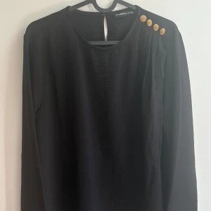 Svart blus med guldknappar  - Stilren svart blus från LC Waikiki med långa ärmar och fyra snygga guldknappar på ena axeln. Blusen har rund halsringning och lätt rynkade detaljer vid axeln för en cool touch. Perfekt för dig som gillar klassisk men trendig stil.
