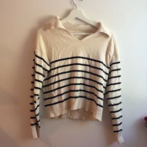 Randig stickad tröja från Gina Tricot - Snygg stickad tröja från Gina Tricot i beige med svarta horisontella ränder. Tröjan har en bred krage, v-ringning och långa ärmar. Ribbstickade muddar vid ärmslut och nederkant ger en extra detalj. Perfekt för dig som gillar en klassisk men trendig look.