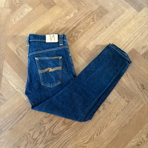 Blå jeans från Nudie Jeans, W30 L28 - Snygga blå jeans från Nudie Jeans med klassiska orange sömmar och ikonisk bakficka med gul brodyr. Modellen har normal passform och raka ben. Tillverkade i slitstark bomull med en mörkblå tvätt. Perfekta för dig som gillar stilrena och tidlösa jeans. Modell Brute Knut 
