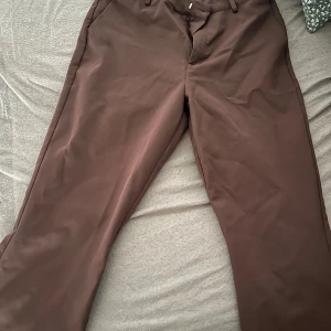 Bruna kostymbyxor från BikBok - Snygga bruna kostymbyxor från BikBok i storlek 42. Byxorna har raka ben, hällor för bälte och klassisk stängning fram. Perfekta för en clean och stilren look, matcha med sneakers eller boots för en trendig vibe. Byxorna är uppsydda några få cm för att passa min längd bättre (är 158cm) men säkert inga problem att sprätta upp om man skulle känna för det.