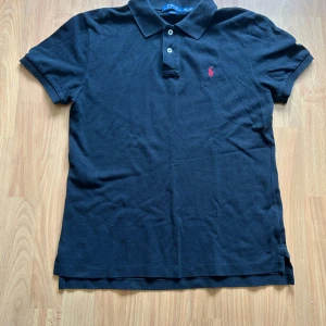 Svart pikétröja Polo Ralph Lauren - Klassisk svart pikétröja från Polo Ralph Lauren med röd broderad logga på bröstet. Slim fit-modell med korta ärmar, krage och två knappar framtill. Tillverkad i mjuk bomull som känns skön mot huden. Perfekt för en clean och stilren look.