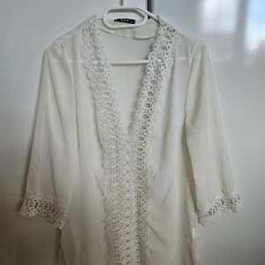 Vit blus med spetsdetaljer från SHEIN - Superfin vit blus från SHEIN med djup v-ringning och breda spetsdetaljer längs halsringningen och ärmsluten. Blusen har trekvartsärm och är gjord i ett lätt, luftigt material som känns fräscht och somrigt.