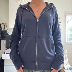 Mörkblå GANT hoodie med dragkedja - Mörkblå ziphoodie från GANT i storlek small. Har klassisk GANT brodering på framsidan och stora framfickor. Är i gott skick, men på insidan vid dragkedjan finns slitningar, dock inget som syns vid användning