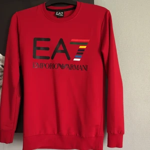Röd EA7 Emporio Armani sweatshirt - Röd sweatshirt från EA7 Emporio Armani med stort tryck framtill i svart och färgglada detaljer på siffran 7. Tröjan har rund hals, långa ärmar och ribbade muddar. Perfekt för dig som gillar streetwear och vill sticka ut med en ikonisk logga.
