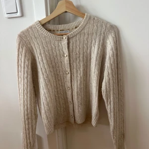 Beige kabelstickad kofta Holly & White - Kabelstickad beige kofta från Holly & White med rund halsringning och pärlemorknappar framtill. 