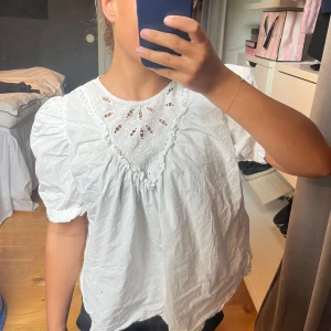 Vit blus med broderad spets - Söt vit blus med kort puffärm och broderad spets upptill. Blusen har en lös passform och fina detaljer med hålbroderi vid halsen. Perfekt för en fräsch och somrig look. Kan behöva strykas för snygghetens skull😅🩷