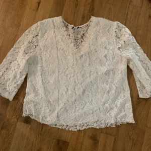 Vit spetsblus med blommönster - Superfin vit blus i spets med blommigt mönster och v-ringning. Blusen har trekvartsärm och är helt genombruten i spets. Perfekt att styla med jeans eller kjol för en söt look. Har klippt längst ner för att den var lite lång förut. Etiketten är borta men ser ut som en storlek S.