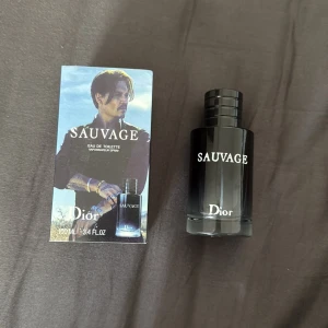 Dior Sauvage Eau de Toilette 100ml - Dior Sauvage Eau de Toilette 100ml med originalförpackning. Flaskan är svart med vit text och har en stilren, rund design. Förpackningen har en bild och tydlig logga. En ikonisk doft som känns modern och fräsch.