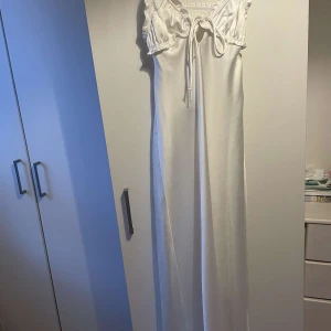 Vit zara långklänning - Supersnygg vit långklänning i satinliknande material med snörning framtill och öppen rygg. Klänningen har volangdetaljer vid axlarna och smala axelband. Klänningen är ifrån zara passar en s/xs
