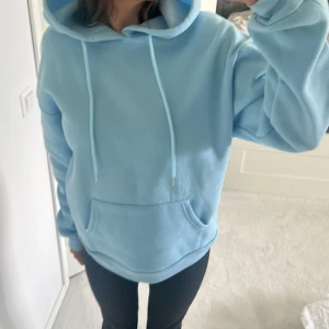 Ljusblå hoodie med tryck bak - Ljusblå hoodie med stor vit text och jordglob-tryck på ryggen. Tröjan har huva med snören, känguruficka framtill och ribbade muddar. Perfekt för chill dagar eller när du vill ha en snygg och bekväm look. Storlek S-M