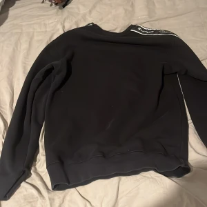 Svart Champion sweatshirt med logga - Svart sweatshirt från Champion i storlek S. Tröjan har vita loggor längs båda ärmarna och en liten broderad Champion-logga på bröstet. Klassisk passform med rund hals och långa ärmar. Perfekt för en sportig och avslappnad stil.