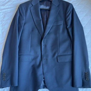 Mörkblå kavaj från These Glory Days - Snygg mörkblå kavaj från These Glory Days med klassisk passform, två knappar framtill och tre fickor. Fodrad insida i blankt tyg och stilrena detaljer. Perfekt för dig som vill ha en stilren och tidlös look.